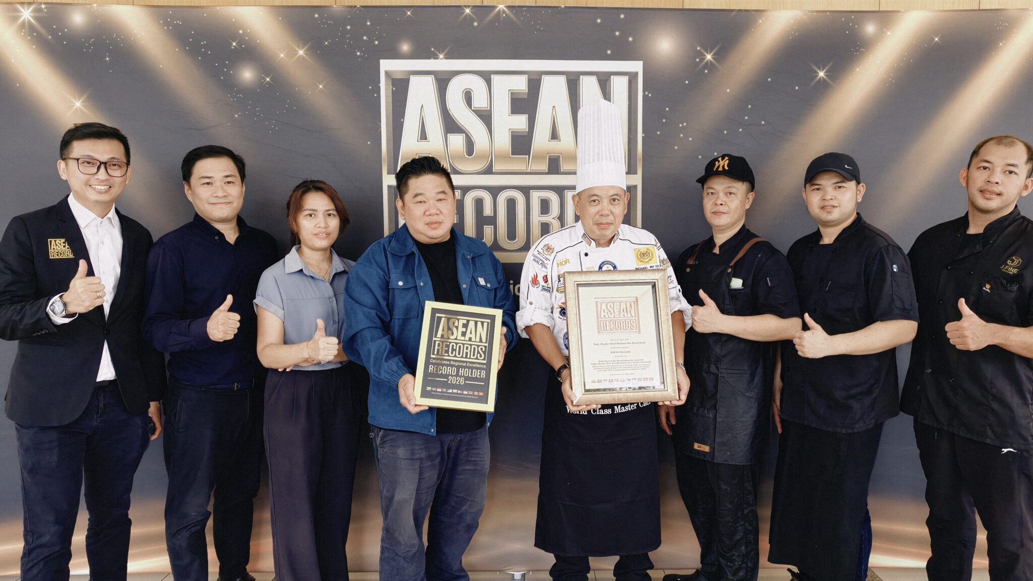 Eight people in dark uniforms pose for a group photo at an ASEAN Records event; center person in a chef's hat holds a framed certificate with others showing thumbs up.