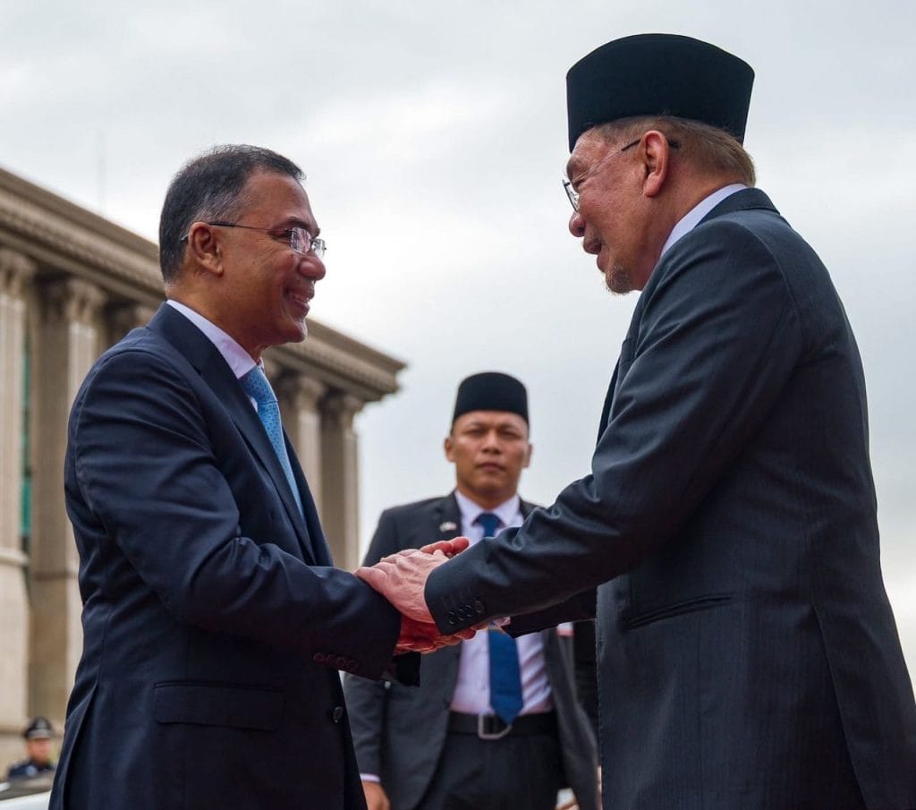 Two men in dark suits shake hands outdoors, one wearing a tall black hat, with a third man in a suit standing behind them.