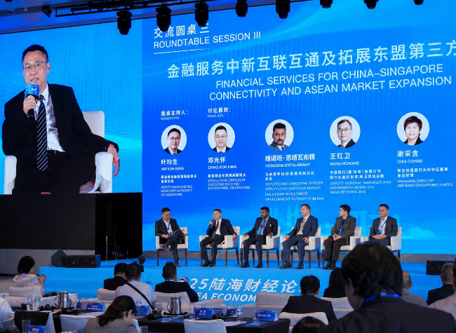 Man speaks into a microphone on a stage with a blue backdrop; panelists sit in chairs as part of a financial services conference discussion, with a large screen displaying speakers' bios in Chinese and English.
