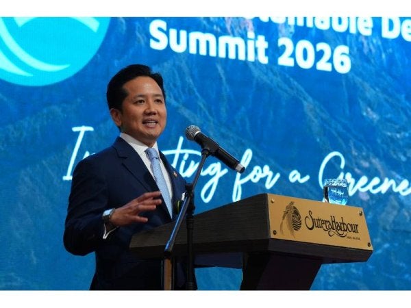 A man in a dark suit giving a speech at a podium on stage with a blue backdrop reading 'Summit 2026' and a microphone