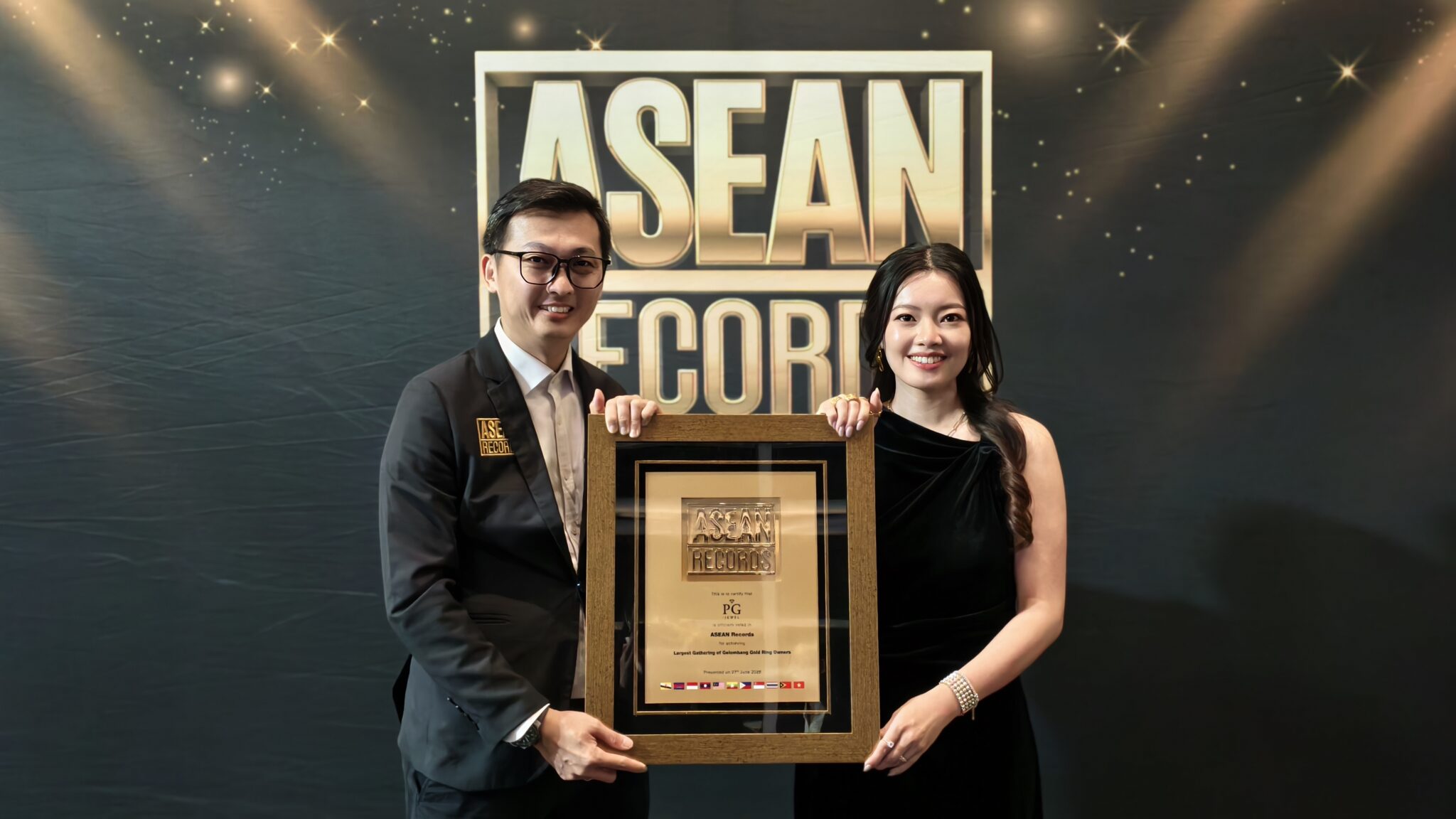 Two formally dressed individuals pose with a large framed award plaque in front of an ASEAN-themed backdrop.