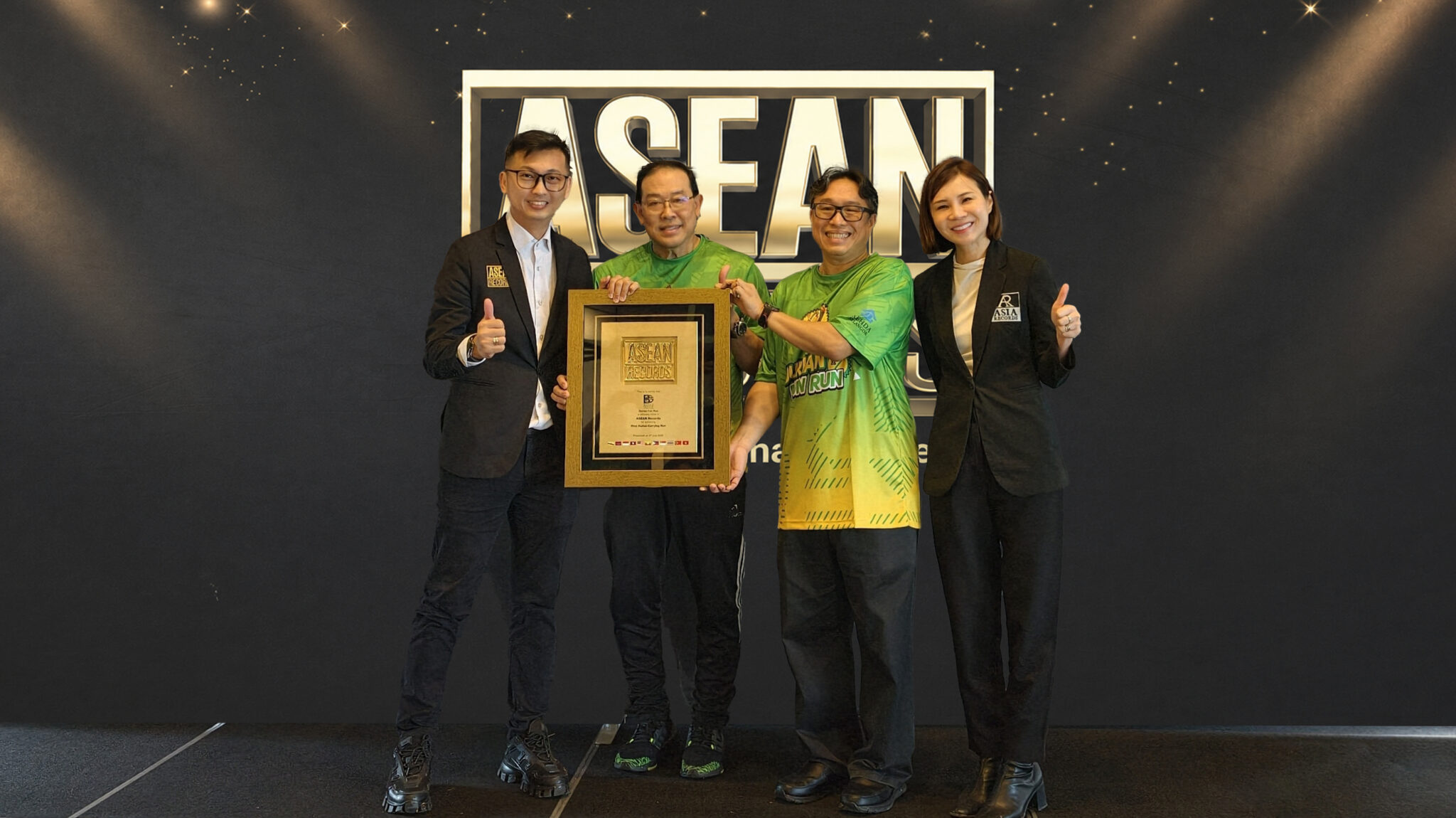 Four people on a stage pose with a framed award plaque in front of a large ASEAN sign, giving thumbs up.