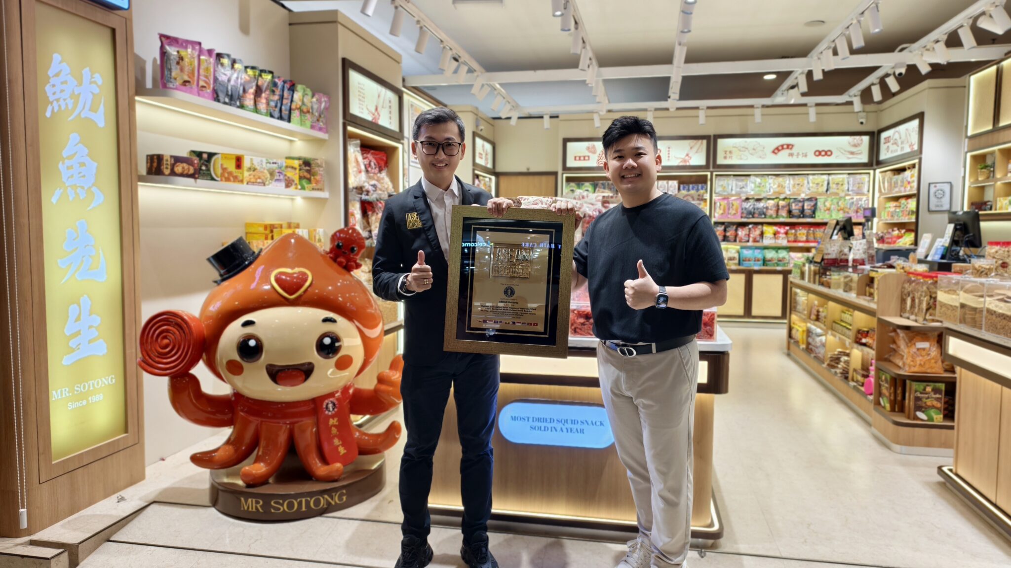 Two men pose with a framed award inside a busy Asian grocery or snack shop, smiling at the camera.