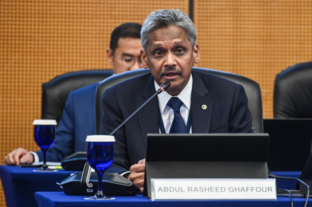 Man in a dark suit and blue tie speaks into a microphone at a formal panel; nameplate reads Abdul Rasheed Ghafour.