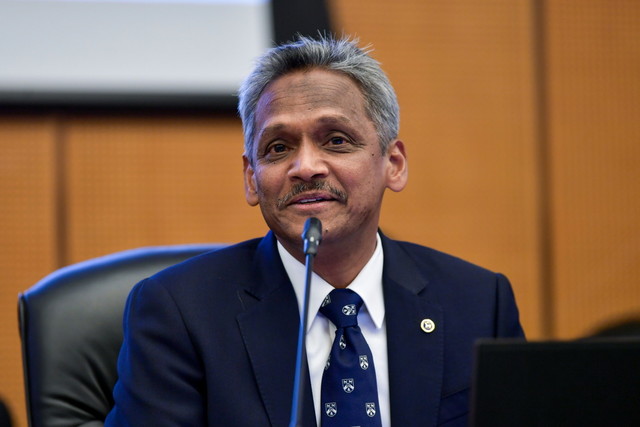 Middle-aged man in a navy suit speaks into a microphone at a formal conference, seated at a panel with wood-paneled walls behind him.