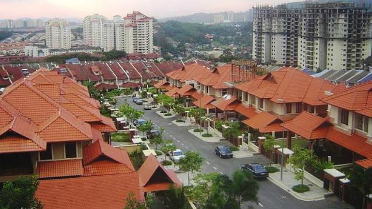 Red-tiled houses form a residential street, with parked cars and green trees, and tall apartment buildings in the background.