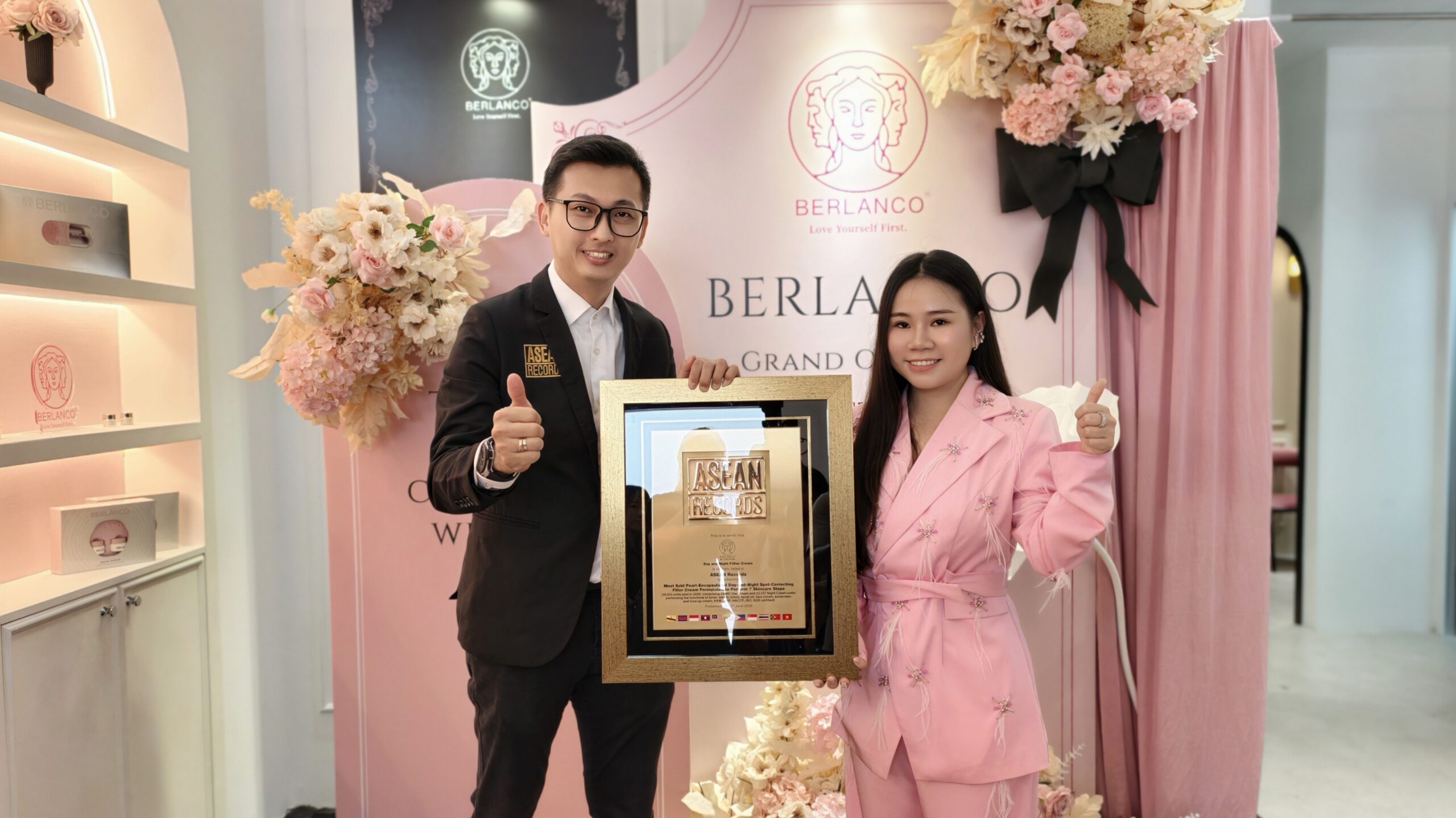 Smiling man in a black suit and woman in a pink suit hold a large framed award together at a pink floral backdrop with BERLANCO branding.