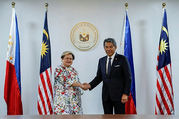 Two officials shake hands in a formal setting, flanked by Philippine and Malaysian flags and a Department of Foreign Affairs seal in the background.
