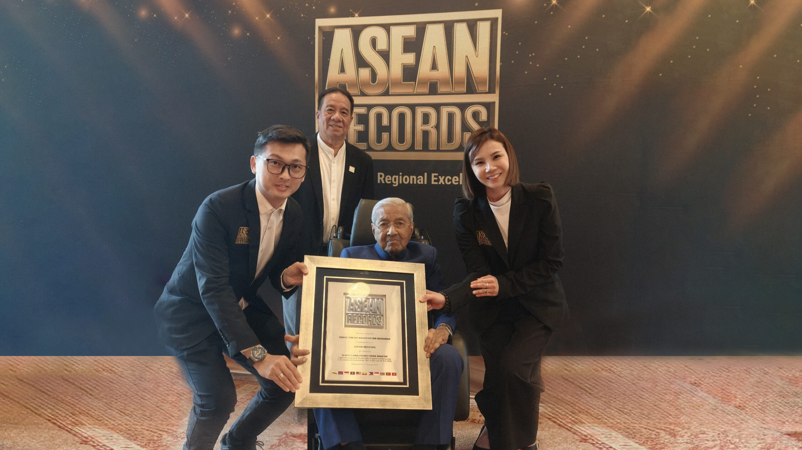 Older man in a wheelchair holding a framed award, flanked by three smiling teammates in suits in front of an ASEAN Records backdrop.