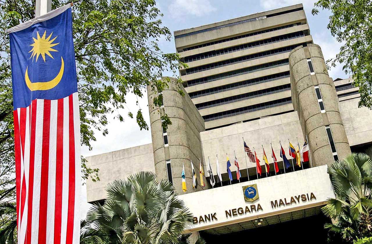 Front view of Bank Negara Malaysia building with a large Malaysian flag in the foreground and a row of international flags on the terrace